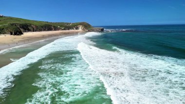 Nefes kesen hava manzaralı gösterişli dalgalar yemyeşil tepelerin çerçevelediği güzel bir sahile çarpıyor.