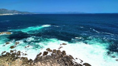 Çarpıcı kıyı manzarası dalgaların, berrak mavi gökyüzünün altındaki engebeli kayalara güçlü bir şekilde çarptığını gösteriyor.
