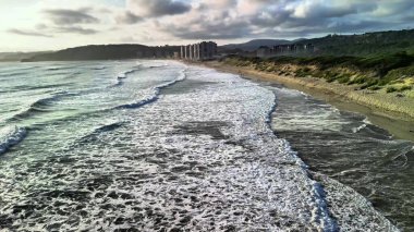Yumuşak okyanus dalgalarının büyüleyici görüntüsü yukarıdaki bulutlu gökyüzünün altındaki kumlu kumsala yavaşça yuvarlanıyor.