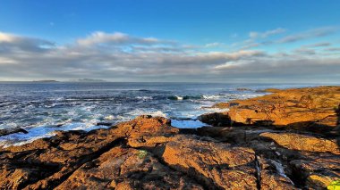 Berrak mavi gökyüzünün altındaki engebeli kayalara çarpan dalgaların çarpıcı görüntüsünü hissedin.