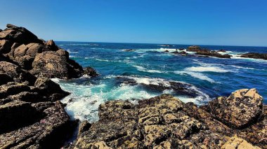 Sakin bir okyanus manzarası, parlak mavi gökyüzünün altında heybetli kayalara çarpan güçlü dalgaları gösterir.