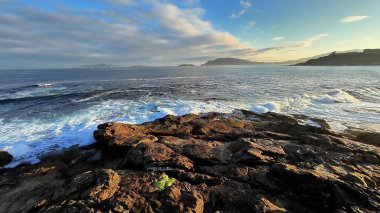 Sakin bir sahil sahnesi, berrak mavi gökyüzünün altında engebeli kayalara çarpan büyüleyici dalgaları gösteriyor.