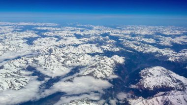 Bu çarpıcı hava görüntüsü, açık mavi gökyüzünün altındaki görkemli karla kaplı dağ zirvelerini güzel bir şekilde vurguluyor.