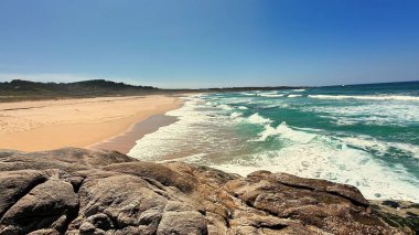 Dalgaların açık mavi gökyüzünün altındaki kayalara çarptığı güzel kumlu bir sahilin nefes kesici manzarası.