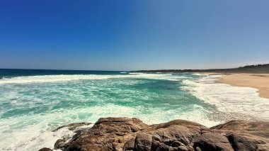 Canlı mavi bir gökyüzünün altında yumuşak dalgalar ve kristal berraklığında sular sergilenen nefes kesici bir sahil manzarası..