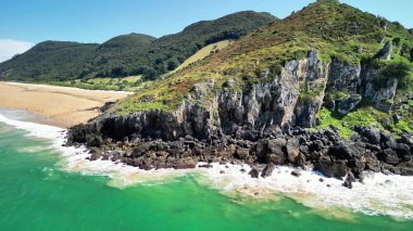 Yeşil tepelerin, kayalık uçurumların ve güneşli bir günde sakin bir plajın nefes kesici manzarası.