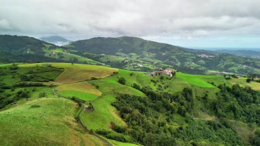 Hava manzarası bulutlu bir gökyüzünün altındaki yemyeşil tepeleri yakalıyor, doğanın dingin güzelliğini sergiliyor.