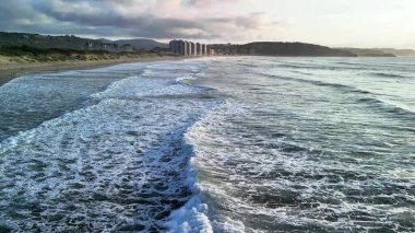 Güzel dalgalar sakin bir plaja çakılırken uzak binalar bulutlu bir gökyüzünün altında duruyor.