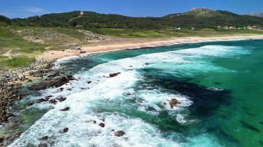 Berrak gökyüzünün altındaki el değmemiş bir sahilde engebeli kayalara çarpan güçlü dalgaların nefes kesici manzarasının tadını çıkar.