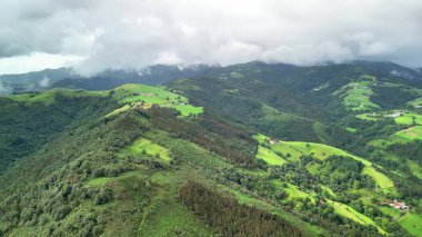 Hava manzarası bulutlu bir gökyüzünün altında doğanın özünü güzel bir şekilde gösteren canlı yeşil tepeleri gösteriyor.