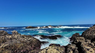 Dalgaların mavi gökyüzünün altında kayalara çarptığı çarpıcı bir sahil manzarası. Plaj keşfi ve rahatlama için mükemmel.