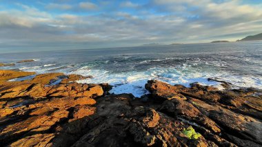 Nefes kesici bir kıyı manzarası dramatik kayalık kıyıları ve bulutlu bir gökyüzünün altında okyanus dalgalarını canlı bir şekilde gösteriyor.