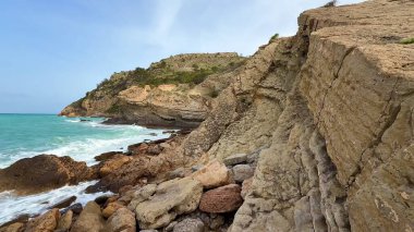 Nefes kesici manzara, canlı turkuaz deniz kıyısında engebeli kayalara çarpan dramatik uçurumları ve dalgaları gösteriyor.