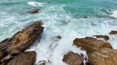 Dalgalar nazikçe çarpışıyor ve engebeli kayalara çarpıyor. Sakin, turkuaz bir sahil şeridi boyunca.