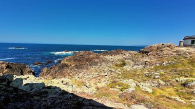 Berrak bir gökyüzünün altında parıldayan mavi okyanusun yanında engebeli, pürüzlü kayaları barındıran nefes kesici bir kıyı manzarası.