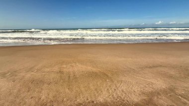 Sakin ve sakin bir sahil sahnesi yumuşak, sıcak kumları sergiliyor ve parlak mavi gökyüzünün altında yavaşça dalgalar süzülüyor.