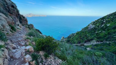 Bereketli yeşillik ve geniş bir okyanus ufkuyla süslenmiş engebeli, kayalık bir sahil yolunun nefes kesici manzarası.