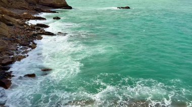 Güzel ve sakin bir sahil manzarası turkuaz suları sergiliyor üzerinde bulutlu bir gökyüzünün altında kayalık uçurumlar var.