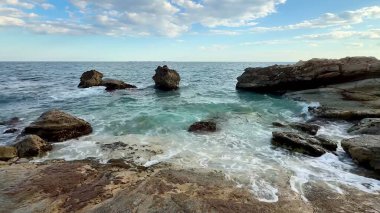 Huzurlu bir sahil sahnesi, bulutlu bir gökyüzünün altında dalgaların engebeli kayalık kıyılara çarptığı.