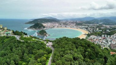 Bir hava fotoğrafında çarpıcı bir plaj, canlı bir şehir manzarası ve ahenkli yemyeşil tepeler sergileniyor.