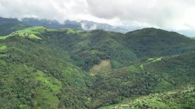 Hava manzarası, dramatik bulutlu bir gökyüzünün altında yemyeşil dağlar ve vadileri gösteriyor. Resimli bir manzara yaratıyor.