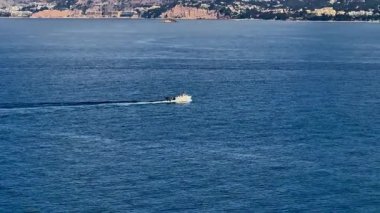 Huzurlu sularda süzülen küçük bir teknenin sakin bir sahnesi. Resimli bir sahil zeminiyle çerçevelenmiş.