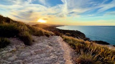 Huzurlu bir patika nefes kesici bir okyanus manzarası sunuyor, gün batımının canlı renklerini görselleştiriyor.