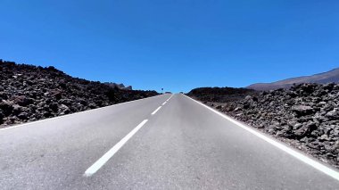 Manzaralı bir asfalt yol, güneşli bir günde çarpıcı manzarayla çerçevelenmiş dramatik bir lav manzarasında esiyor.