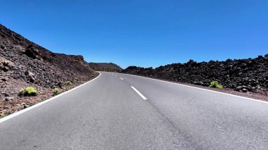 Eşsiz volkanik kaya oluşumlarıyla çevrili uzun, dolambaçlı bir yol, açık mavi bir gökyüzünün altında uzanır.