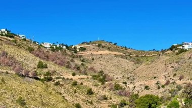 Göz kamaştırıcı bir yamaç manzarası, parlak, açık mavi gökyüzünün altında, izleyiciyi büyüleyen lüks evler barındırır.
