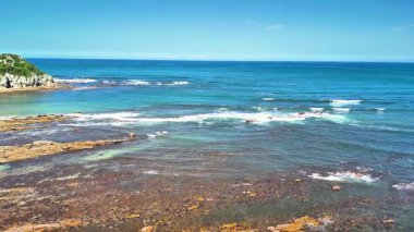Güzel bir okyanus manzarası, yumuşak dalgalar kayalık bir kıyı şeridini aydınlatıyor, parlak güneş ışığıyla aydınlanıyor.