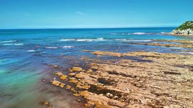 Çarpıcı bir okyanus manzarası parlak mavi gökyüzünün altında ince bulutlu kayalık bir kıyı şeridi ve berrak bir su gösteriyor.