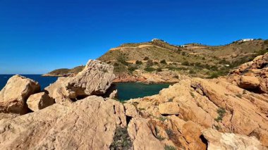Berrak mavi gökyüzünün altında kayalık uçurumlar ve sakin okyanus sularıyla nefes kesici kıyı şeridi manzarası.