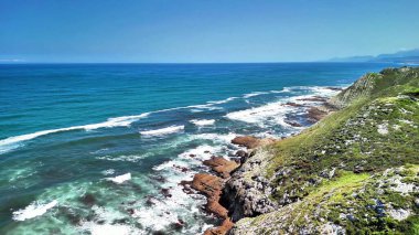 Zümrüt kayalıkların ve açık mavi gökyüzünün altında güçlü dalgaların olduğu nefes kesici bir kıyı şeridi manzarası