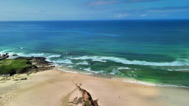 Yumuşak dalgalar ve yemyeşil manzaralarla donatılmış huzurlu bir plajı güzel bir şekilde gösteren dinamik bir manzara.