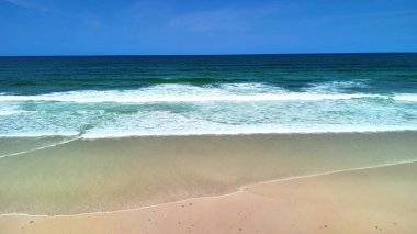 Okyanus dalgalarının nefes kesici görüntüsü çarpıcı bir şekilde kıyıya çarpıyor canlı, parlak mavi gökyüzünün altında