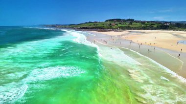 Canlı dalgaların ve canlı yeşil suların tadını çıkaran çeşitli insanlarla dolu parlak ve güneşli bir sahil sahnesi.
