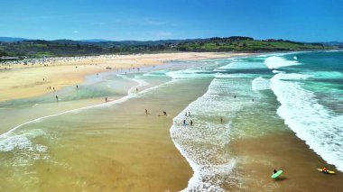 Parlak bir sahil, yeşil tepeler ve güneşli gökyüzünün altında dalgalanan dalgaların çarpıcı hava manzarasını tecrübe edin.