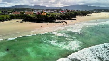 Çarpıcı bir sahil manzarası var. Canlı dalgalar, kumlu bir kıyı şeridi ve çevresindeki manzaralı tepeler.