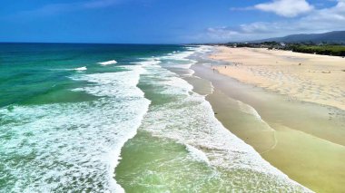 Nefes kesen bir hava manzarası, sakin bir kumsalın sakin dalgaları ve güneşli kumları yansıtır.