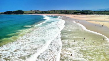 Sörfçülerin, altın kumun ve canlı dalgaların bulunduğu çarpıcı bir sahil manzarası, hepsi açık bir gökyüzünün altında.