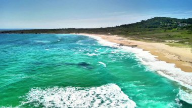 Çarpıcı bir hava manzarası, canlı turkuaz sular ve yumuşak okyanus dalgalarıyla süslenmiş sakin bir plajı gösteriyor.