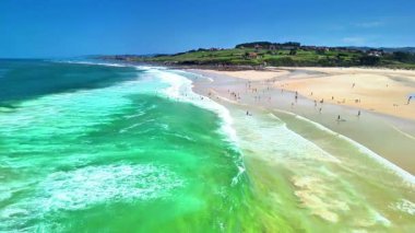 Canlı dalgaların ve canlı yeşil suların tadını çıkaran çeşitli insanlarla dolu parlak ve güneşli bir sahil sahnesi.