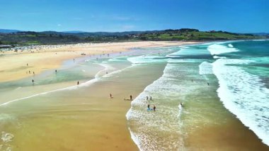 Parlak bir sahil, yeşil tepeler ve güneşli gökyüzünün altında dalgalanan dalgaların çarpıcı hava manzarasını tecrübe edin.