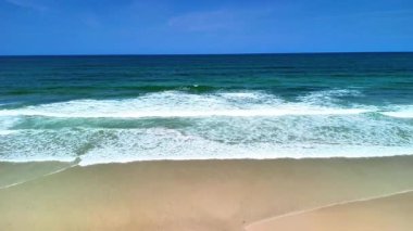 Okyanus dalgalarının nefes kesici görüntüsü çarpıcı bir şekilde kıyıya çarpıyor canlı, parlak mavi gökyüzünün altında