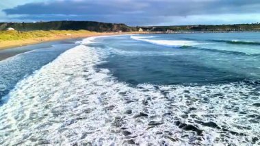Okyanus dalgalarının nefes kesici görüntüsü canlı bir günbatımı gökyüzünün altındaki kumlu sahile nazikçe çarpıyor.