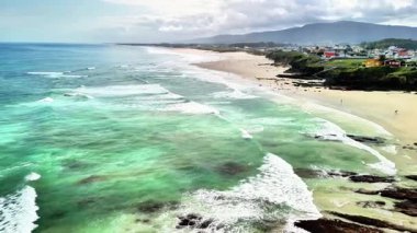 Hava manzarası, canlı dalgalar ve yakınlardaki büyüleyici kıyı evleri ile işaretlenmiş güzel, sakin bir sahili yakalar.