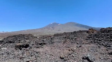 Yüksek bir volkanın nefes kesen ve çarpıcı manzarası ve parlak mavi gökyüzünün altındaki engebeli lav arazisi.