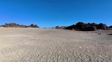 Göz kamaştırıcı ve geniş bir çöl manzarası, yukarıdaki parlak mavi gökyüzünün altında kayalık oluşumları vurguluyor.