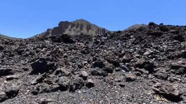 Açık mavi gökyüzünün altındaki kayalık araziyi gösteren büyük volkanik bir manzaranın nefes kesici ve çarpıcı görüntüsü.